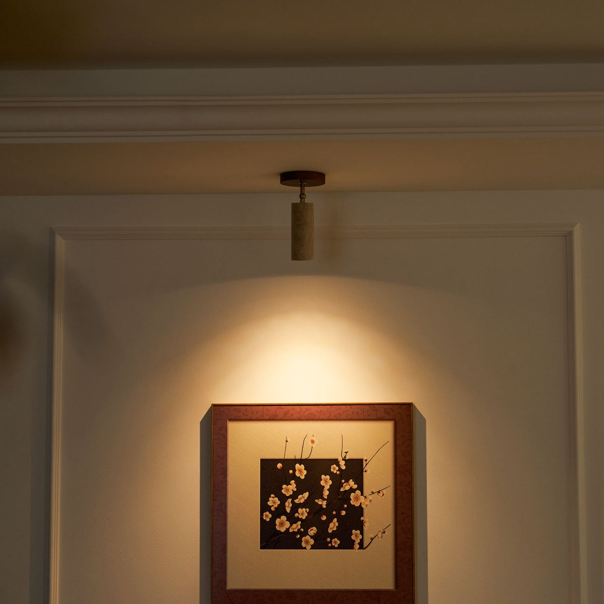Lit Clérachic travertine spotlight in a dim room, casting a warm pool of light over a framed blossom print on a panelled wall.