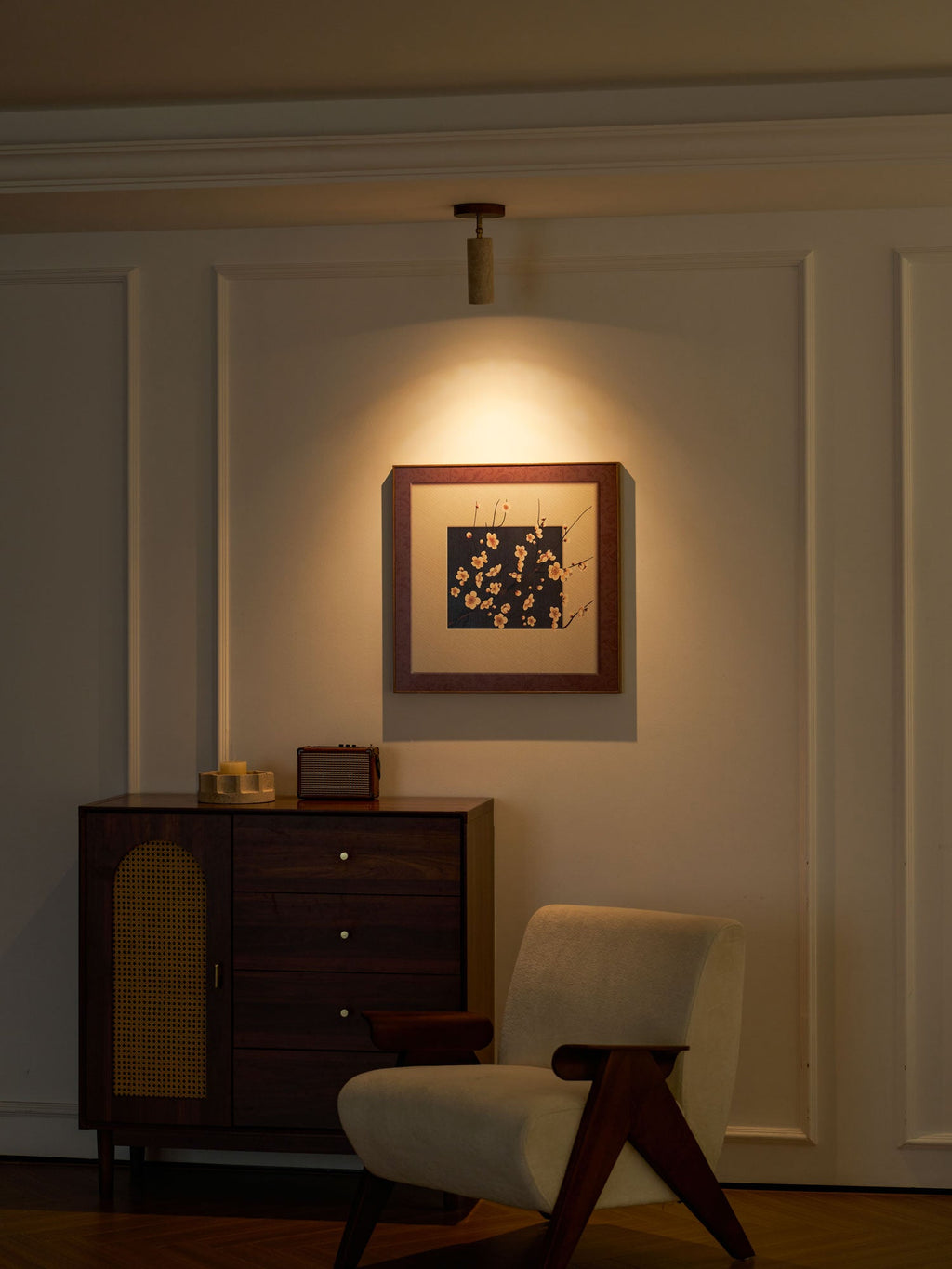 Evening room view of the Clérachic spotlight lighting a framed print, with a walnut cabinet and a cream lounge chair on a wood floor.