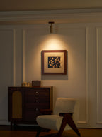 Evening room view of the Clérachic spotlight lighting a framed print, with a walnut cabinet and a cream lounge chair on a wood floor.