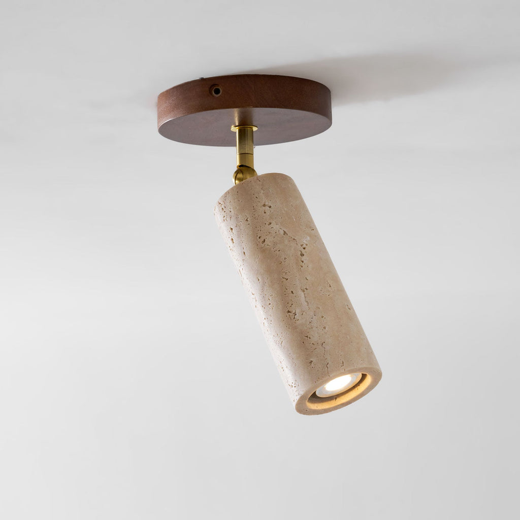 Looking up at this Clérachic spotlight on a white ceiling, a walnut plate, brass pivot and travertine cylinder with the lamp lit.