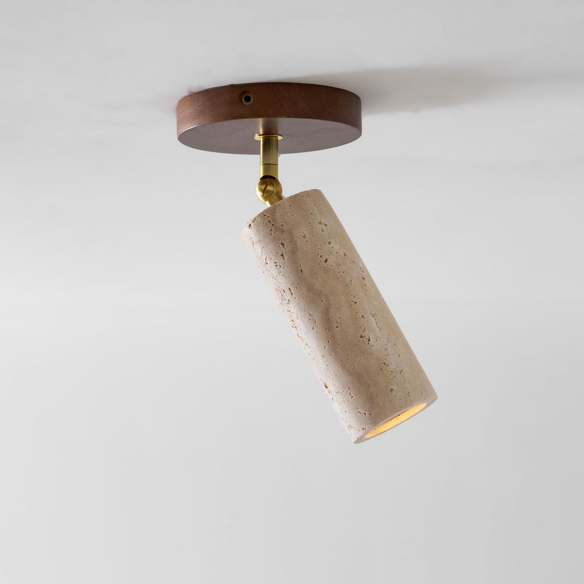 Clérachic travertine spotlight tilted on a brass joint from a round walnut ceiling plate, shown against a pale ceiling.