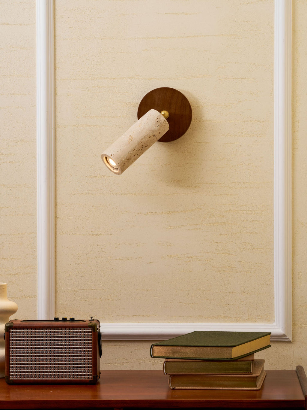 Wall-mounted Clérachic spotlight turned to the left on a cream wall, with a retro speaker and a pile of books on a walnut surface.