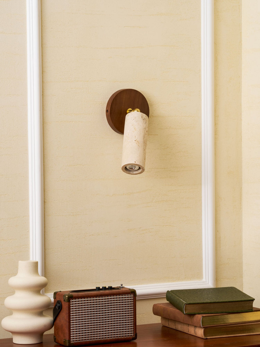 Clérachic travertine spotlight pointing down from a walnut wall plate on a cream wall, with a vase, a speaker and books below.