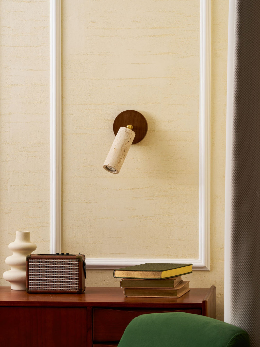 Smaller view of the wall-mounted Clérachic travertine spotlight on a cream panelled wall above a speaker, a vase and a green chair.