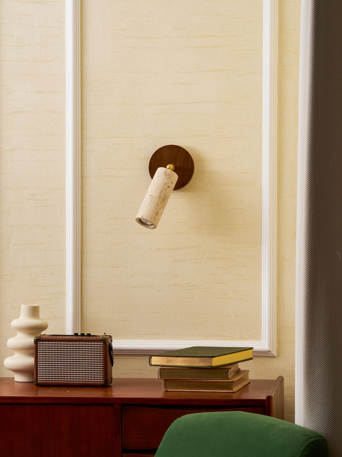 Smaller view of the wall-mounted Clérachic travertine spotlight on a cream panelled wall above a speaker, a vase and a green chair.