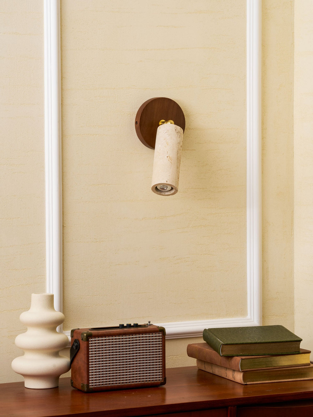 Clérachic travertine spotlight aimed down on a walnut wall plate, above a cream vase, a retro speaker and a stack of green books.