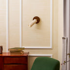 Clérachic wall spotlight pointing left on a cream panelled wall, with a green armchair, a walnut drawer unit and books below.
