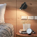 Black Clérachic reading light on a wood-panelled wall, its head angled down and glowing over a bedside table with flowers and a clock.