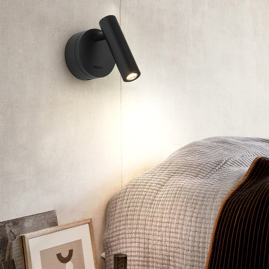 Beside a bed with a knitted throw, this Clérachic black spotlight sits on a cream wall, its head tilted down to cast a soft pool of light.