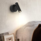 Beside a bed with a knitted throw, this Clérachic black spotlight sits on a cream wall, its head tilted down to cast a soft pool of light.