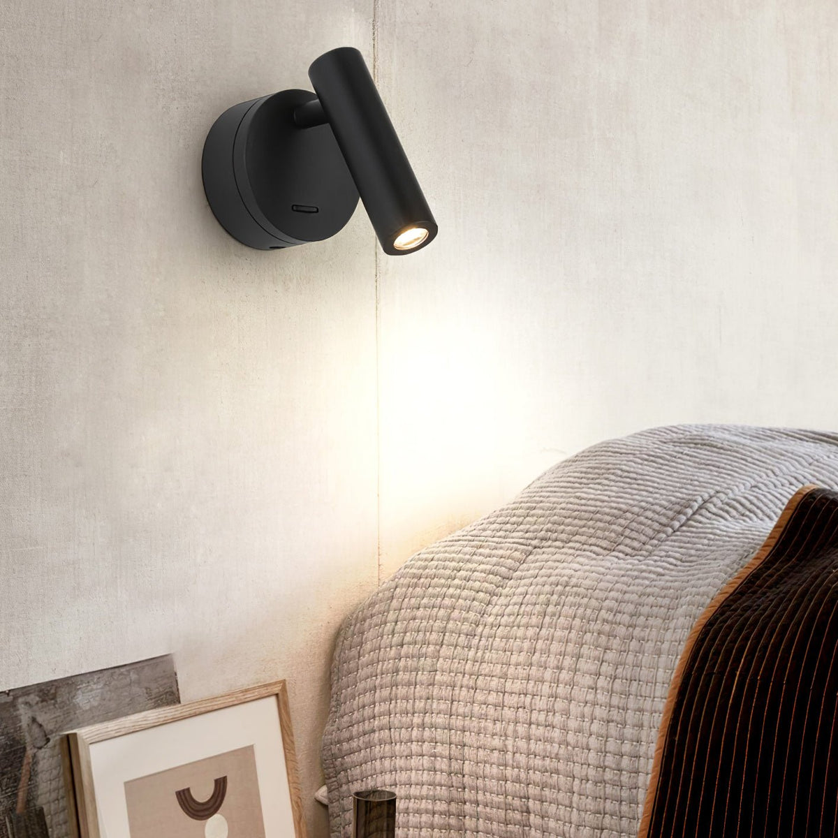 Beside a bed with a knitted throw, this Clérachic black spotlight sits on a cream wall, its head tilted down to cast a soft pool of light.