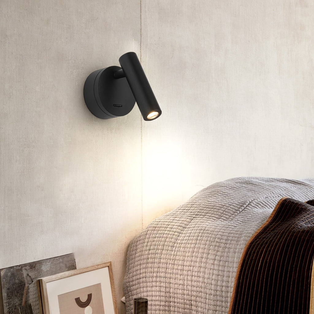 This Clérachic reading light in black, with its cylinder head angled up and to the side, lights a cream wall above a knitted bed throw.