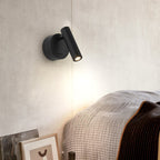 This Clérachic reading light in black, with its cylinder head angled up and to the side, lights a cream wall above a knitted bed throw.