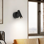 A black Clérachic spotlight wall light on a pale wall beside a tall window, with the arm of a chair and a tan cushion below.