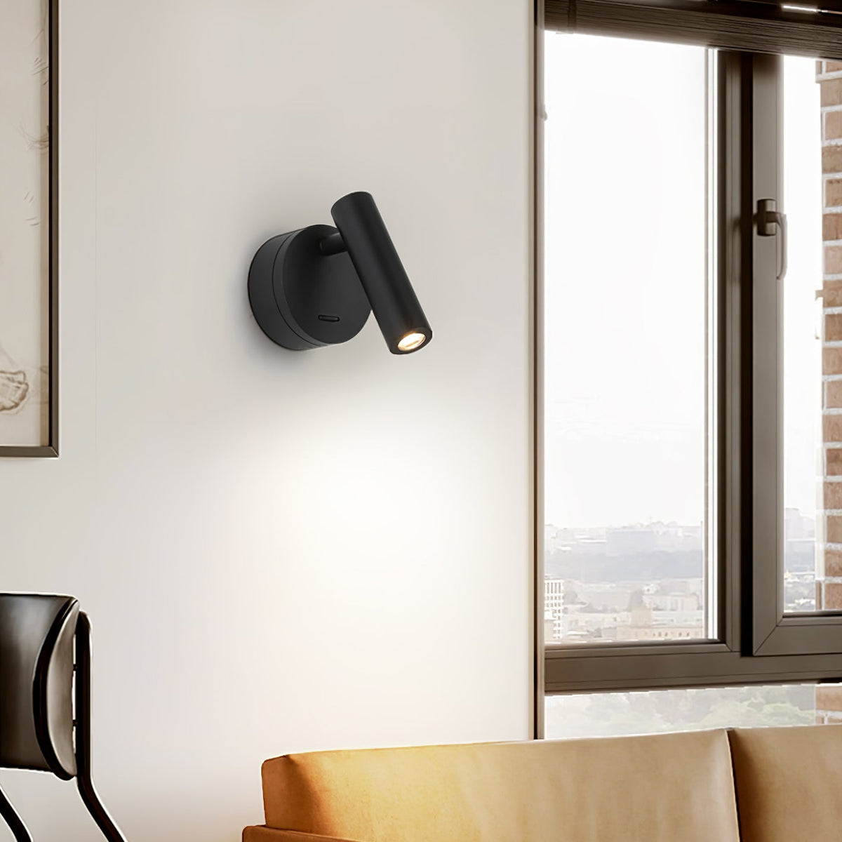 A black Clérachic spotlight wall light on a pale wall beside a tall window, with the arm of a chair and a tan cushion below.