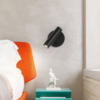 Black Clérachic wall light above a teal bedside table and an orange upholstered headboard, its head turned down and to the side.