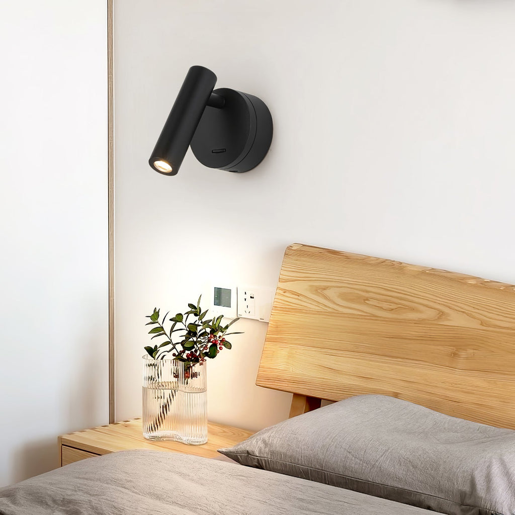 A black Clérachic wall light beside a pale wooden headboard, lit softly above a bedside table with a vase of berries and leaves.