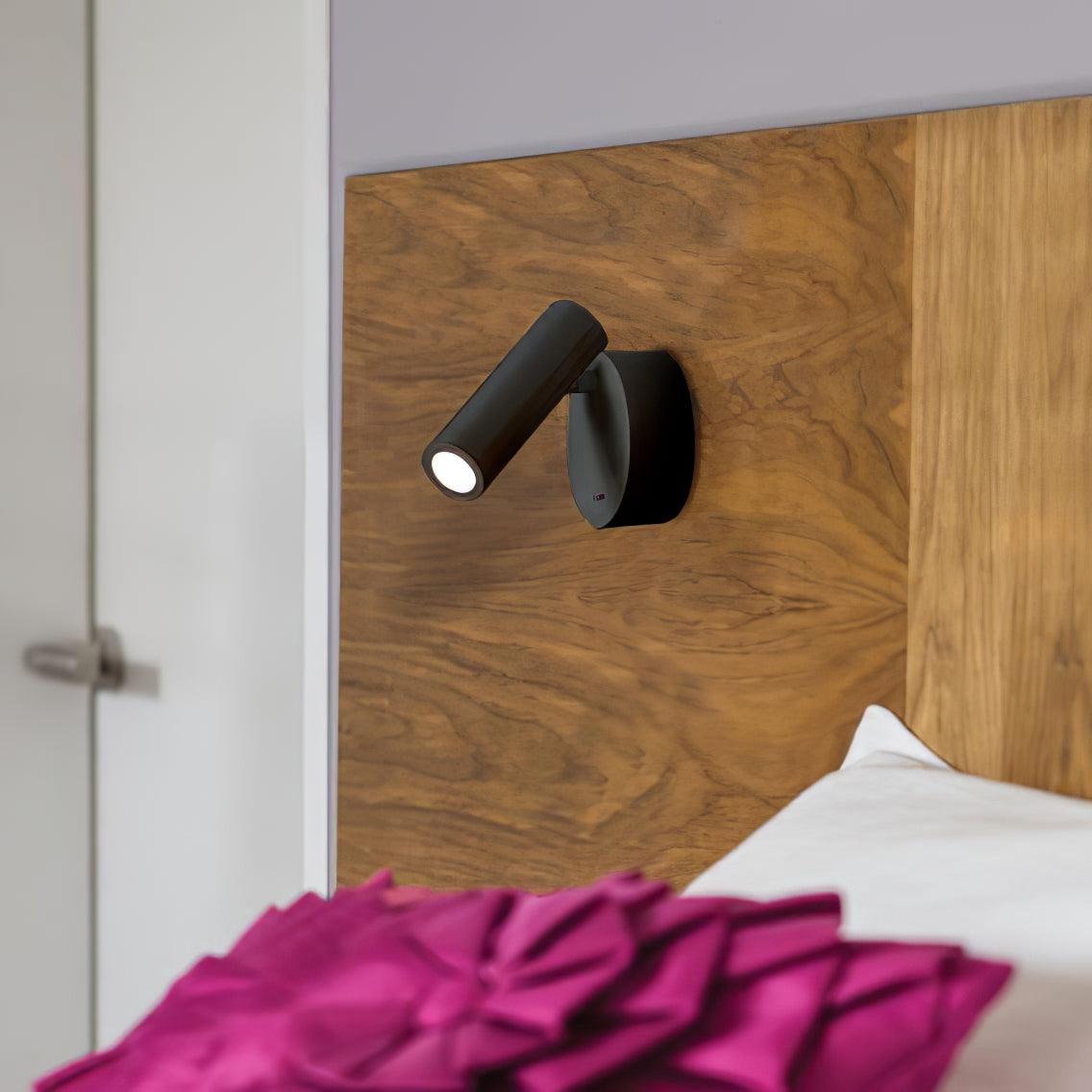 Small black Clérachic spotlight wall light on a walnut-veneer headboard panel, with a magenta cushion at the foot of the frame.