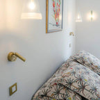 A gold-toned Clérachic wall light beside a bed with a palm-print cover, with a pair of glass pendants and a framed print above.