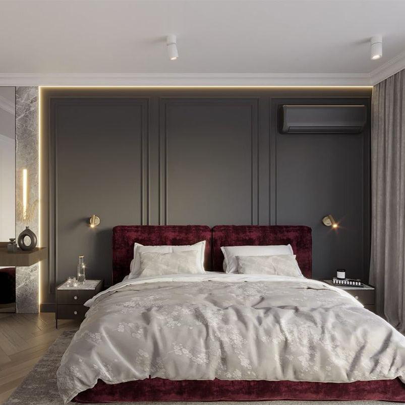 Two small gold-toned Clérachic wall lights sit on either side of a dark panelled headboard wall, above a bed with a burgundy headboard.