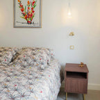 This Clérachic wall light, in a gold tone, on a pale wall above a bed with palm-print bedding and a walnut-toned bedside cabinet.