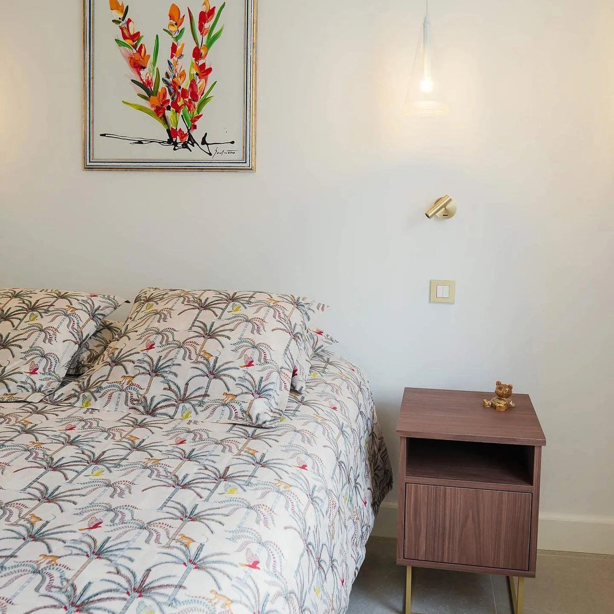 This Clérachic wall light, in a gold tone, on a pale wall above a bed with palm-print bedding and a walnut-toned bedside cabinet.