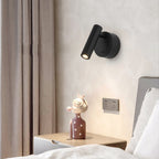 A black Clérachic reading light on a cream wall beside curtains, above a bedside table with a figurine and a bed pillow.