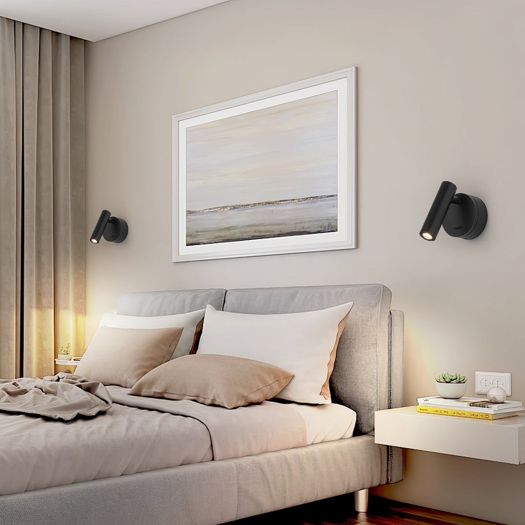 Two black Clérachic spotlight wall lights hang either side of a framed seascape above a grey upholstered bed with a warm glow behind it.