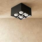 Clérachic black square box ceiling downlight with four glowing round spots and white reflector cells, studio shot on a limewash ceiling.
