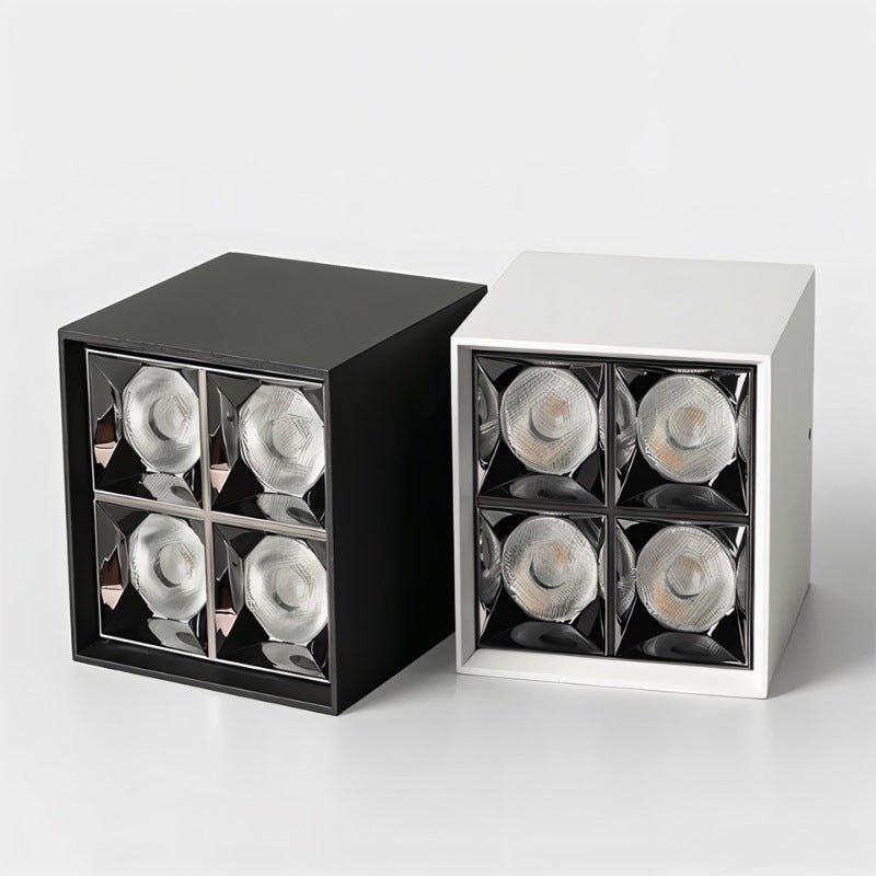 Close up of a black and a white Clérachic square box downlight, each showing four faceted reflector cells, on a light grey studio backdrop.