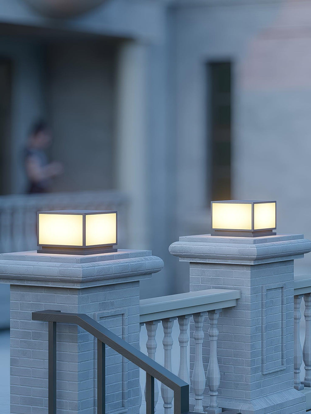Close view of two lit Clérachic post lights on brick pillars, with a stone balustrade and a handrail beside the steps.