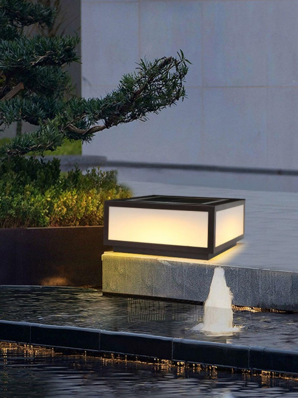 Single Clérachic post light lit on a low stone ledge beside a small water feature, with a pine branch above it.