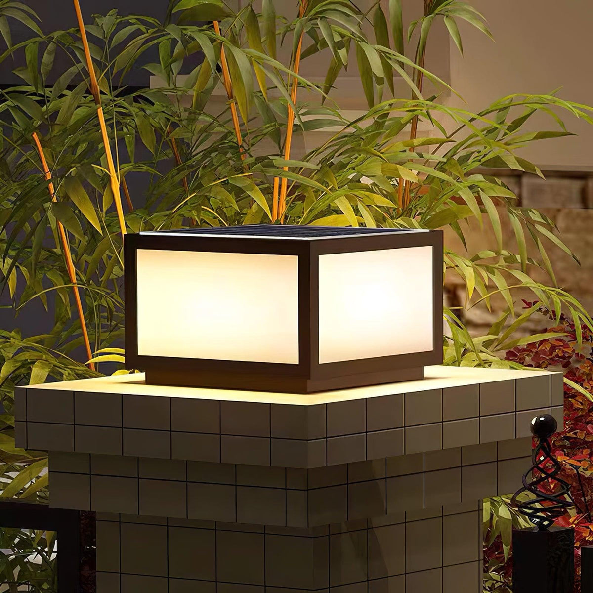 Lit Clérachic square post light on a tiled pillar, surrounded by bamboo leaves, with a soft glow under its base.