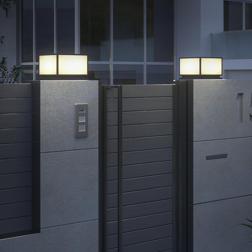 Lit square Clérachic post lights on grey pillars beside a horizontal-slatted gate, with a modern building behind.