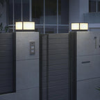 Lit square Clérachic post lights on grey pillars beside a horizontal-slatted gate, with a modern building behind.