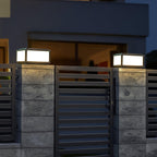 Two lit Clérachic post lights on stone-clad pillars either side of a black slatted gate, with a house window glowing behind.