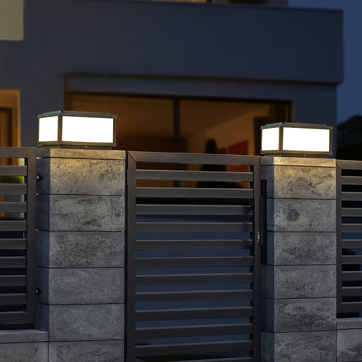 Two lit Clérachic post lights on stone-clad pillars either side of a black slatted gate, with a house window glowing behind.