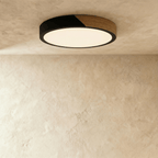 Clérachic round flush ceiling light in black and wood, glowing softly against a textured plaster ceiling above a bare wall.