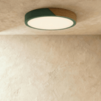 Clérachic round flush ceiling light in deep green and wood, lit warm, with the glow washing across the plaster around it.