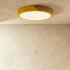 Clérachic round flush ceiling light in golden yellow and wood, lit warm, the yellow band bright against the pale plaster.