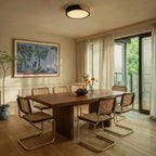 Clérachic round flush ceiling light in black and wood, glowing above a dark wood dining table ringed with cane-backed chairs.