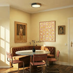 Clérachic round flush ceiling light in black and wood, set on the ceiling above a tufted banquette dining nook lined with framed pictures.