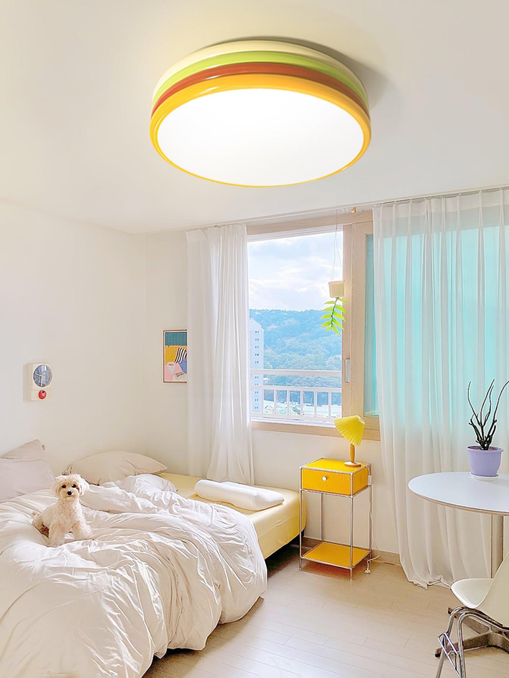 Clérachic stacked ceiling light glowing above a white bedroom with a dog on the bed, a yellow bedside unit and a sheer curtain.