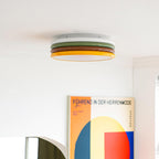 Clérachic ceiling light with its white, green, red-brown and yellow tiers, above a large geometric poster in a white room.