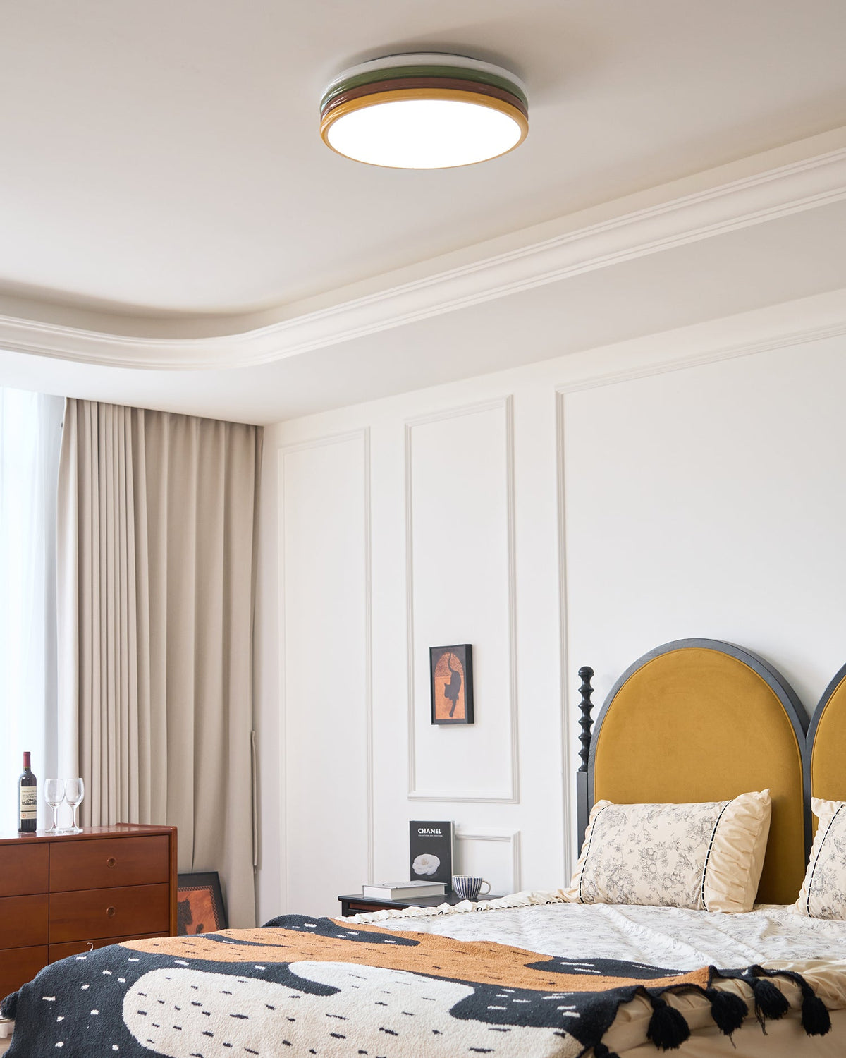 Clérachic ceiling light lit above a bedroom with a mustard arched headboard, a patterned throw and a wood chest of drawers.