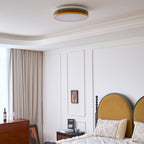 Small Clérachic ceiling light overhead in a white panelled bedroom, with beige curtains, a chest of drawers and a mustard headboard.