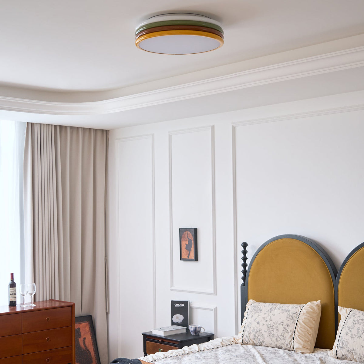 Small Clérachic ceiling light overhead in a white panelled bedroom, with beige curtains, a chest of drawers and a mustard headboard.