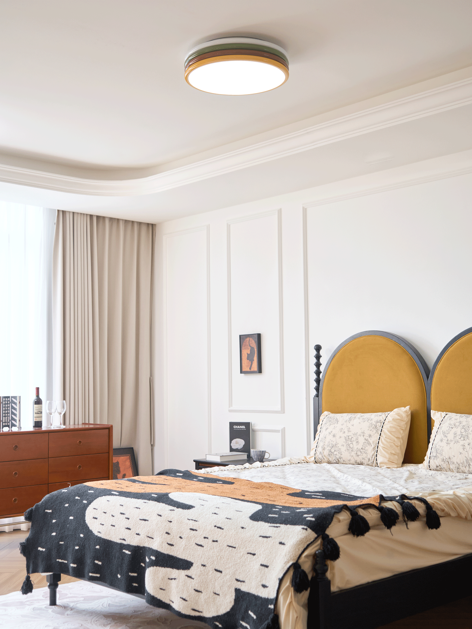 Clérachic ceiling light lit above a bed with a mustard headboard and a black and cream patterned throw with tassels.