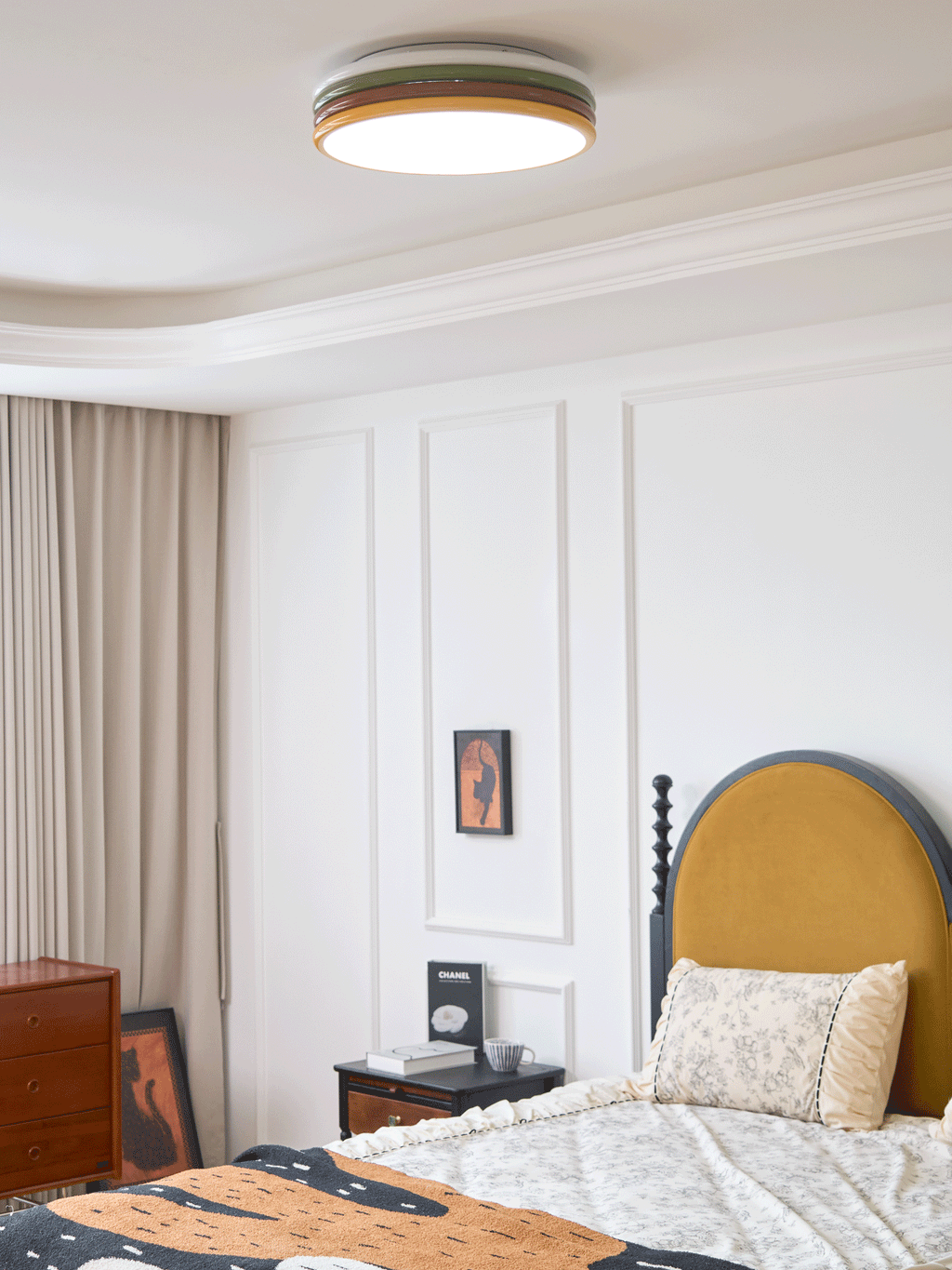 Lit Clérachic ceiling light above a bed with a floral cushion, a mustard headboard and a framed print by the nightstand.