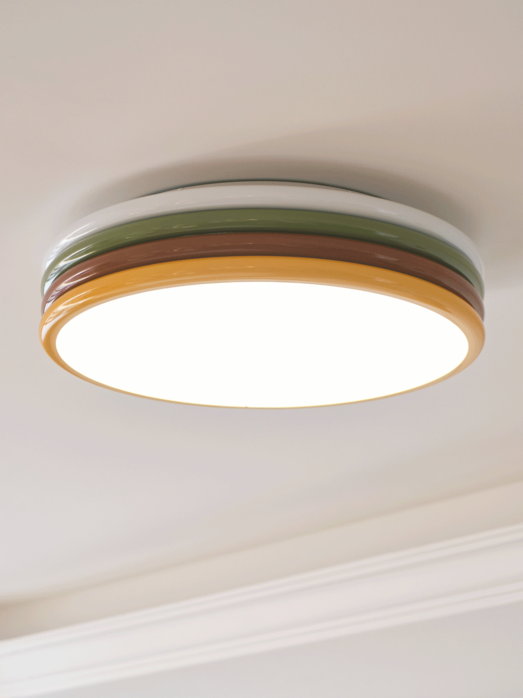 Lit Clérachic ceiling light with a bright diffuser and thin yellow rim, seen from below beside a white cornice.